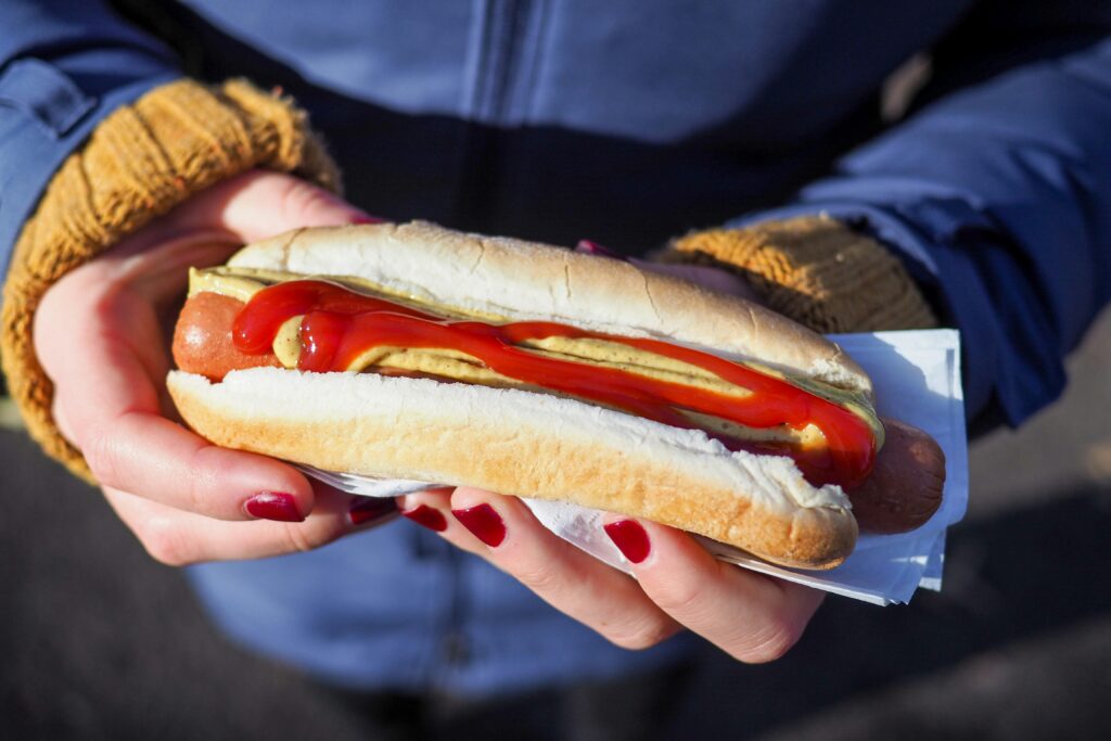 A person with painted nails and full sleeved clothing holds a hot dog with ketchup and mustard.