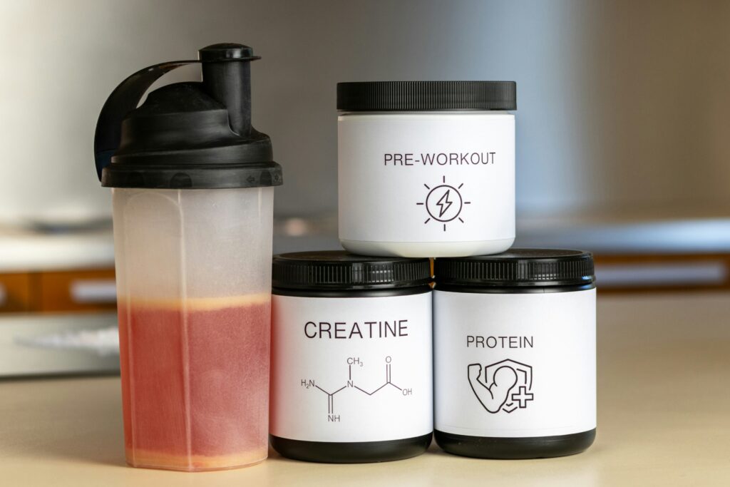 Three containers of dietary and workout supplements stacked beside a blender-bottle with protein shake.