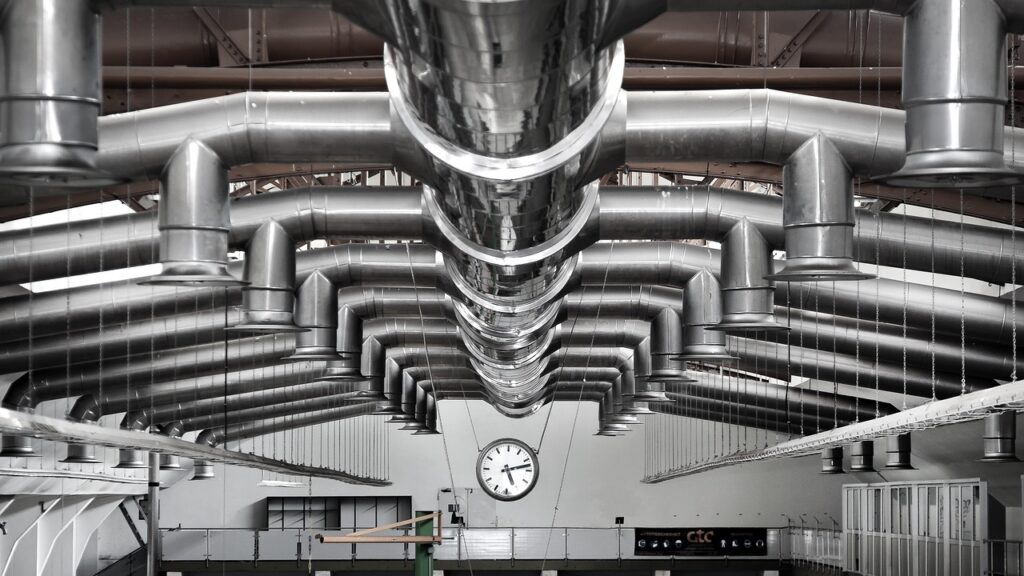 ChatGPT said: The image shows a large industrial facility with an extensive network of metallic air ducts and ventilation systems suspended from the ceiling. At the center is a prominent wall clock, suggesting a structured, timed process environment. This setup is characteristic of automated testing or production lines, potentially used in glass and ceramic manufacturing for quality control, material stress testing, or thermal processing. The symmetrical design and clean layout reflect a highly controlled industrial atmosphere.