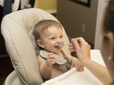 An adult feeds baby food with a spoon to a smiling baby in a high chair.