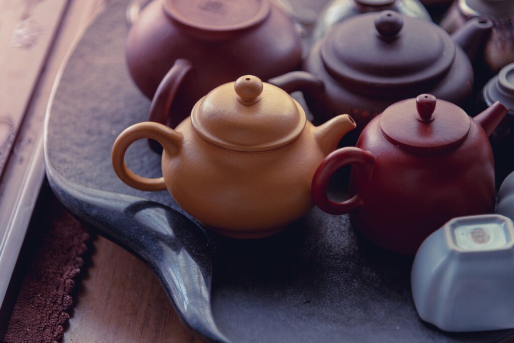 The image features a collection of traditional ceramic teapots displayed on a textured black tray. The teapots vary in shape and color, including shades of beige, red, brown, and dark gray, with a smooth, matte finish typical of handcrafted pottery. The warm lighting enhances their earthy tones, highlighting the craftsmanship and natural aesthetics of the ceramic material. This setup suggests a focus on artisanal ceramics, possibly for testing hardness, glaze quality, or showcasing design in a tea culture context