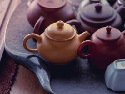 The image features a collection of traditional ceramic teapots displayed on a textured black tray. The teapots vary in shape and color, including shades of beige, red, brown, and dark gray, with a smooth, matte finish typical of handcrafted pottery. The warm lighting enhances their earthy tones, highlighting the craftsmanship and natural aesthetics of the ceramic material. This setup suggests a focus on artisanal ceramics, possibly for testing hardness, glaze quality, or showcasing design in a tea culture context