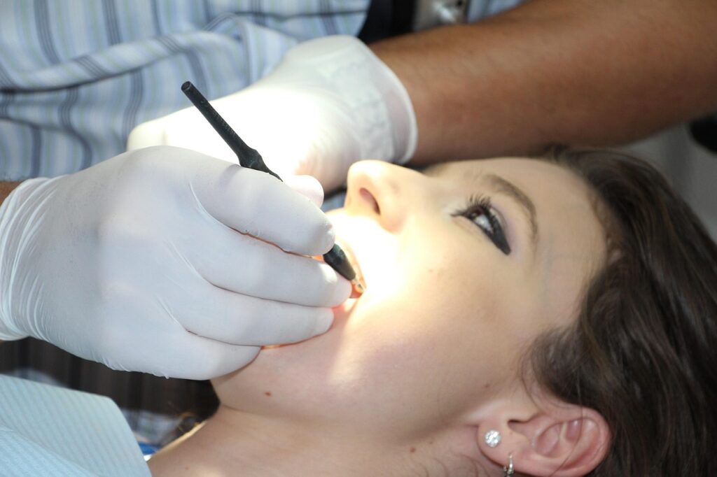 The image shows a close-up scene of a dental procedure in progress. A patient is lying back with their mouth open, while a dentist—wearing white gloves—uses a dental tool, likely for examination or treatment. The brightly lit oral cavity suggests use of focused lighting for visibility. This context implies the application of ceramic materials in medical implants, such as dental crowns, bridges, or fillings, which are valued for their biocompatibility, durability, and aesthetic resemblance to natural teeth.
