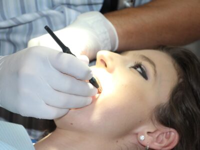 The image shows a close-up scene of a dental procedure in progress. A patient is lying back with their mouth open, while a dentist—wearing white gloves—uses a dental tool, likely for examination or treatment. The brightly lit oral cavity suggests use of focused lighting for visibility. This context implies the application of ceramic materials in medical implants, such as dental crowns, bridges, or fillings, which are valued for their biocompatibility, durability, and aesthetic resemblance to natural teeth.