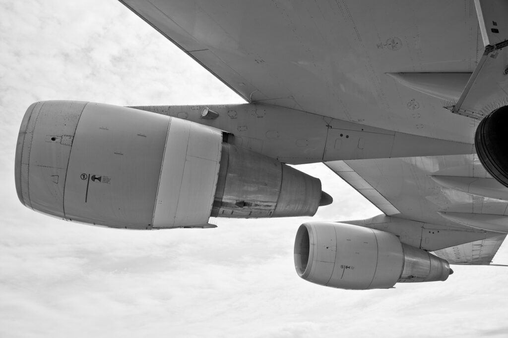 The image shows a close-up, underside view of a large aircraft wing and its engines in black and white. Visible are multiple jet turbines, detailing the nacelles and structural components of the engine mounts. This context highlights the use of ceramics in aerospace engineering, particularly in thermal shielding, engine components, and high-stress zones. Advanced ceramics are commonly utilized here for their high heat resistance, lightweight properties, and durability, which are essential in the demanding environments of aviation and aerospace systems.