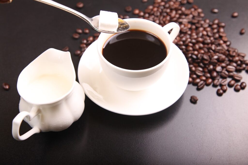 Cup of black coffee on a white saucer with a sugar cube on a spoon above it, next to a small pitcher of cream and scattered coffee beans.