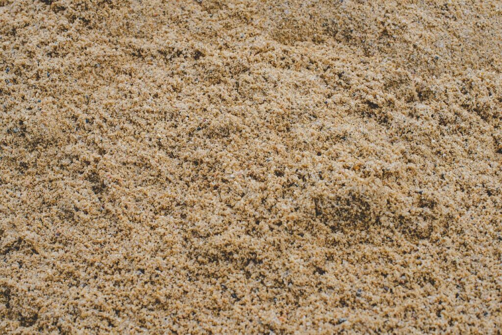 The image shows a close-up view of fine, light brown sand or granular material, likely used as a raw component in the manufacturing or analysis of glass and ceramics. The texture is uniform with small, angular grains, indicating high silica content—essential for glass production. This type of material is typically involved in compositional analysis, where elemental or mineral makeup is examined to ensure quality and performance in glass or ceramic products.