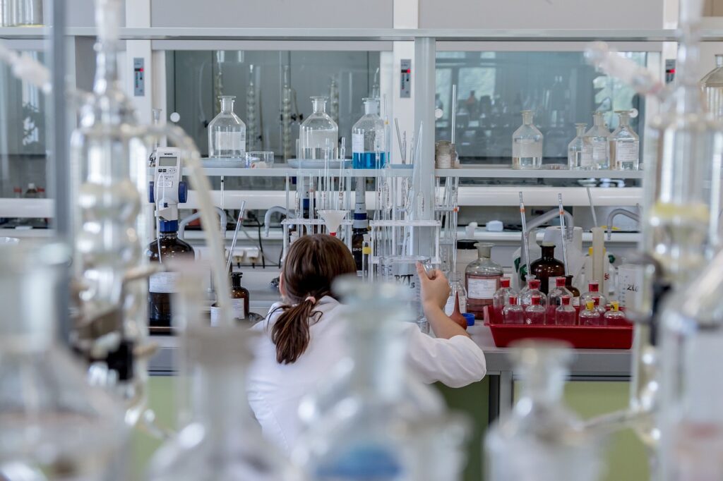 The image shows a scientist in a white lab coat working in a well-equipped laboratory. The individual is seated at a bench surrounded by an array of glassware including beakers, flasks, burettes, and reagent bottles. The setting reflects a controlled environment dedicated to custom test program development, possibly for chemical analysis, material testing, or quality control. The variety of equipment suggests the lab supports flexible experimental setups tailored to specific research or product validation needs.