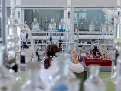 The image shows a scientist in a white lab coat working in a well-equipped laboratory. The individual is seated at a bench surrounded by an array of glassware including beakers, flasks, burettes, and reagent bottles. The setting reflects a controlled environment dedicated to custom test program development, possibly for chemical analysis, material testing, or quality control. The variety of equipment suggests the lab supports flexible experimental setups tailored to specific research or product validation needs.