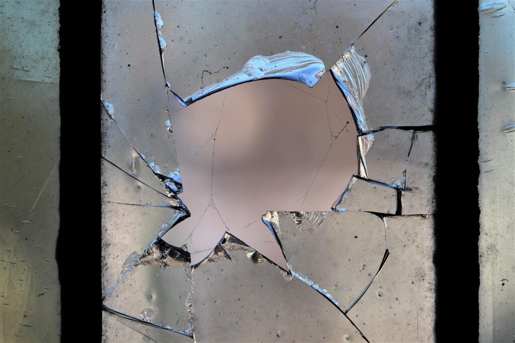 The image shows a pane of broken glass with a large, jagged hole in the center and sharp radial cracks extending outward. The damage pattern indicates impact stress, likely from a forceful blow. The edges of the break are uneven and fractured, showing typical characteristics of brittle materials like glass or ceramics under mechanical failure. This scene is likely related to impact resistance testing or documentation of breakage behavior in safety or materials research.