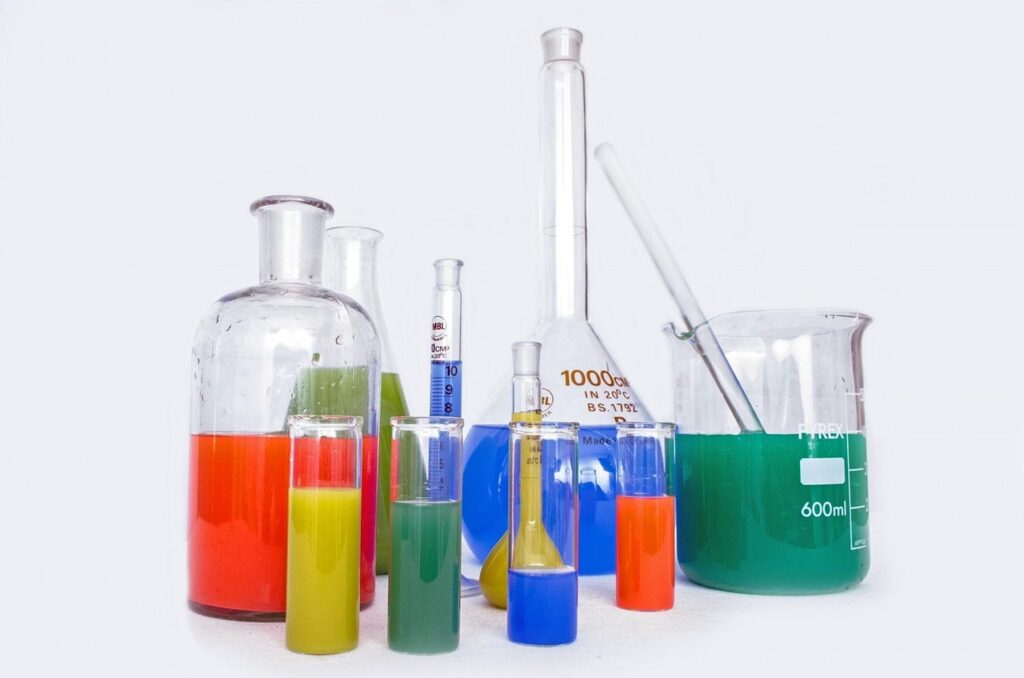 he image displays a variety of laboratory glassware filled with vividly colored chemical solutions. Included are beakers, graduated cylinders, flasks (including a 1000 ml volumetric flask), and test tubes. The liquids are in multiple colors—red, green, yellow, blue, and orange—suggesting different chemical substances or pH indicators. A glass stirring rod rests inside the 600 ml beaker on the right. The setup appears to be used for chemical experiments, material testing, or educational demonstrations involving glass and ceramic materials.