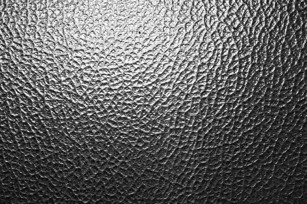 The image shows a close-up of a textured surface that resembles a fine, uneven pattern of cracks or folds, often associated with delamination in materials like laminated glass or coated ceramics. The varying light reflections emphasize the roughness and separation of layers, which could indicate internal stress, adhesive failure, or environmental degradation. This pattern is characteristic in studies of material fatigue, structural failure, or quality control for advanced glass or ceramic composites.