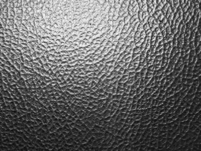 The image shows a close-up of a textured surface that resembles a fine, uneven pattern of cracks or folds, often associated with delamination in materials like laminated glass or coated ceramics. The varying light reflections emphasize the roughness and separation of layers, which could indicate internal stress, adhesive failure, or environmental degradation. This pattern is characteristic in studies of material fatigue, structural failure, or quality control for advanced glass or ceramic composites.
