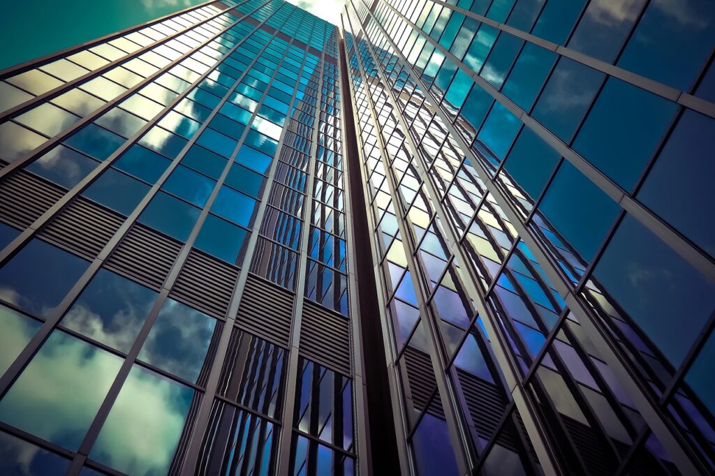 The image shows a dramatic upward perspective of a modern glass skyscraper. The building’s exterior is covered in reflective blue-tinted glass panels, mirroring the sky and clouds, which adds to its sleek and contemporary appearance. The strong vertical lines and symmetry create a sense of height and architectural sophistication, emphasizing the building's towering presence.