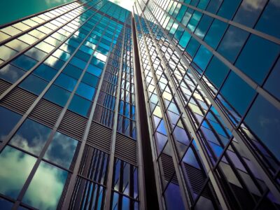 The image shows a dramatic upward perspective of a modern glass skyscraper. The building’s exterior is covered in reflective blue-tinted glass panels, mirroring the sky and clouds, which adds to its sleek and contemporary appearance. The strong vertical lines and symmetry create a sense of height and architectural sophistication, emphasizing the building's towering presence.