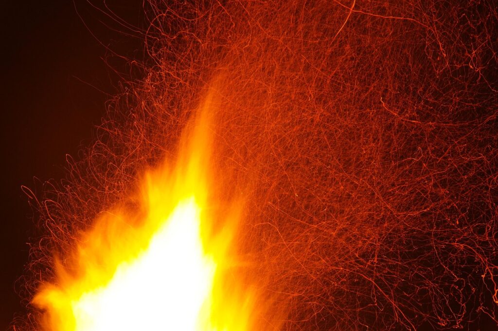The image captures a vivid scene of intense flames and flying sparks against a dark background, suggesting a high-temperature environment—likely associated with thermal testing or fire resistance evaluation. The bright yellow and orange flames dominate the left side, while swirling red embers or particles trail across the right, indicating dynamic heat movement. This visual could represent thermal stress testing of materials like glass or ceramics to observe their behavior under extreme heat conditions.