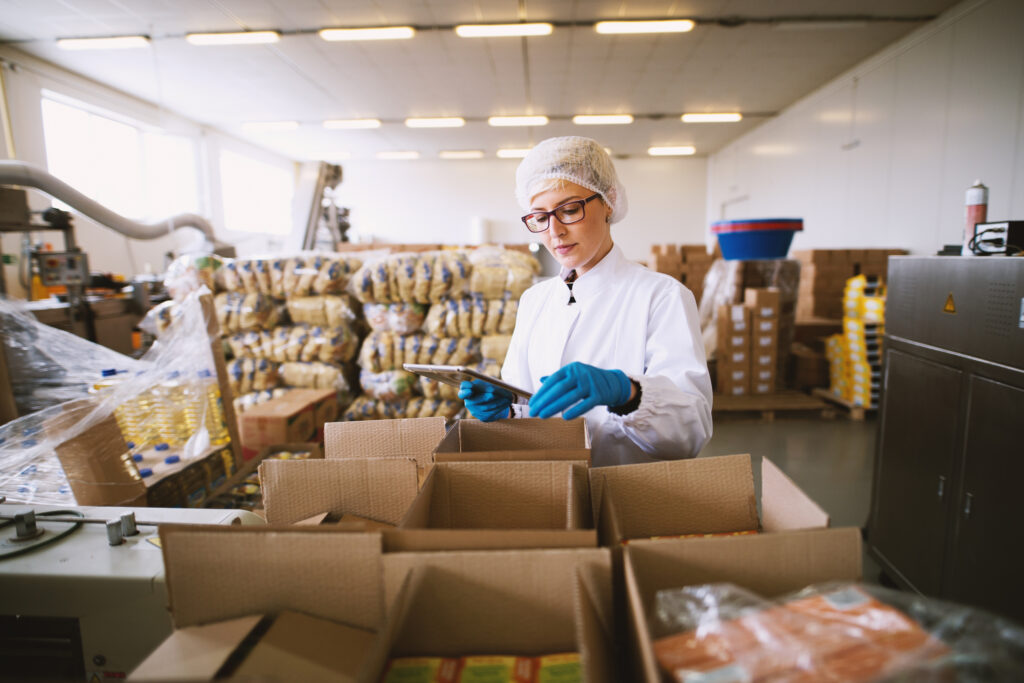Young female worker in PPE is using tablet to check right number of consumer products packages.