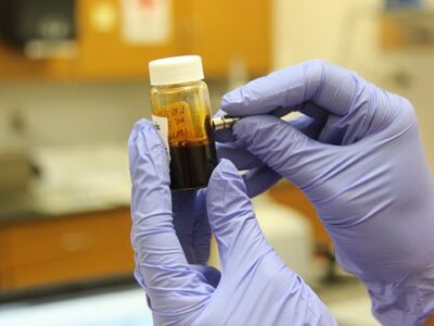 Gloved hands examine a glass bottle filled with a dark liquid.