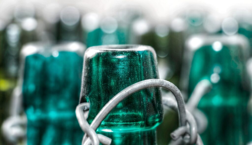 The image shows a close-up of green glass bottle tops with metal swing-top closures. The focus is on the smooth, reflective surface of one bottle, while others blur into the background. This scene highlights sustainable glass packaging, emphasizing reusability, recyclability, and durability—core features in eco-friendly design for beverages and other consumables.