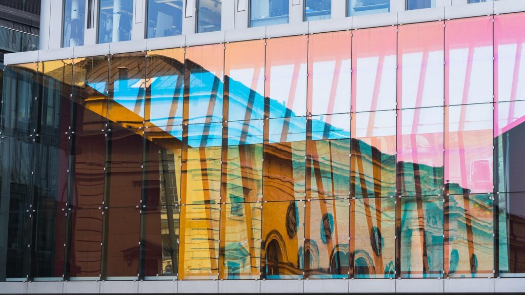The image shows colorful architectural glass panels reflecting a historic building. The glass likely includes optical coatings or laminates, tested for solar control, UV filtering, impact resistance, and aesthetic reflection, common in modern building design.